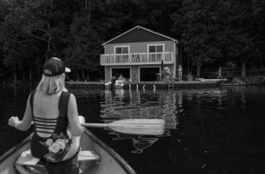 Woman paddling a canoe towards a lakeside house, capturing summer relaxation.